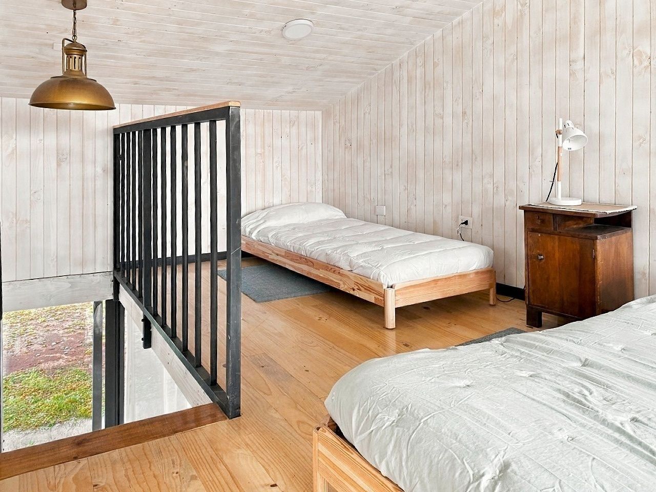 A bedroom with two beds, a wooden floor, a black metal bed frame, a white bedside table, and a hanging light fixture.