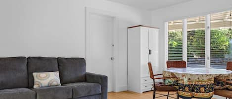 Een kamer met een grijze bank, een eettafel met stoelen en een raam met jaloezieën.