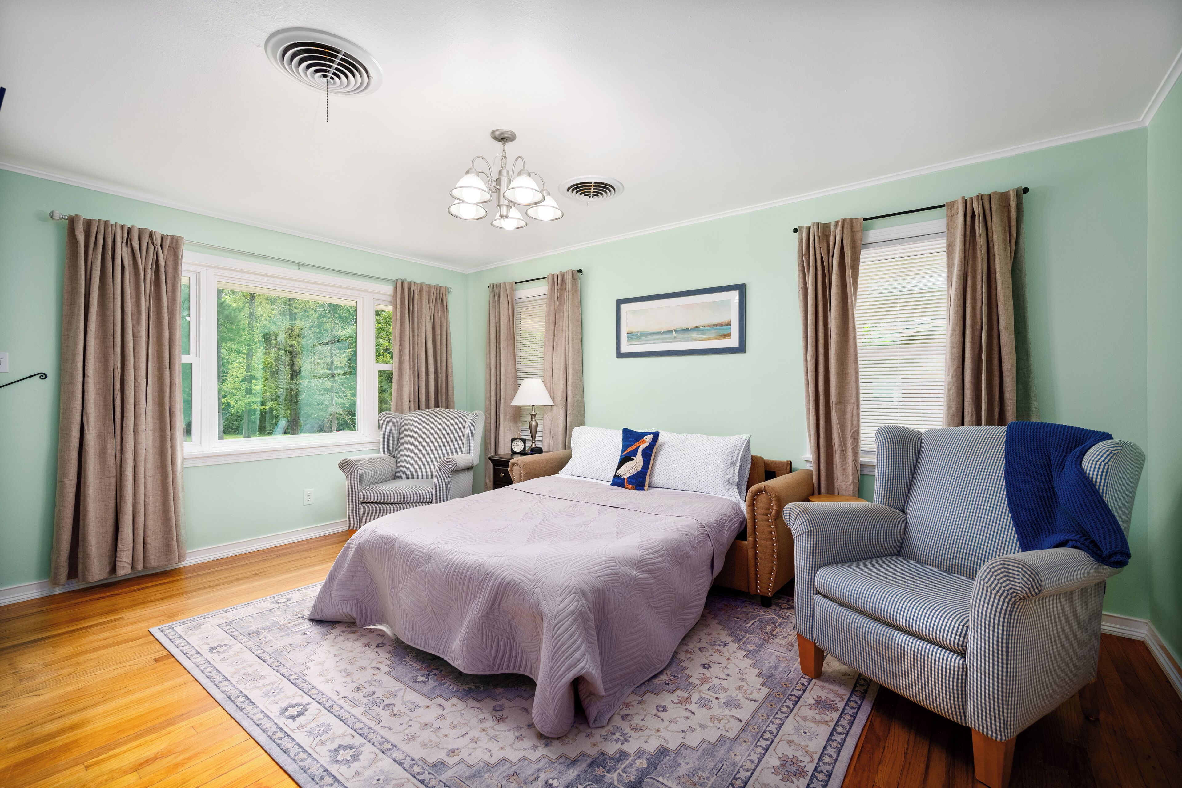 Une chambre à coucher avec un lit, deux chaises, une fenêtre avec des rideaux, un tableau au mur et un ventilateur de plafond.