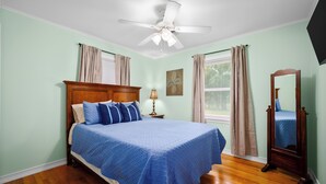 A bedroom with a bed, a ceiling fan, a nightstand, a lamp, a window with curtains, and a mirror.