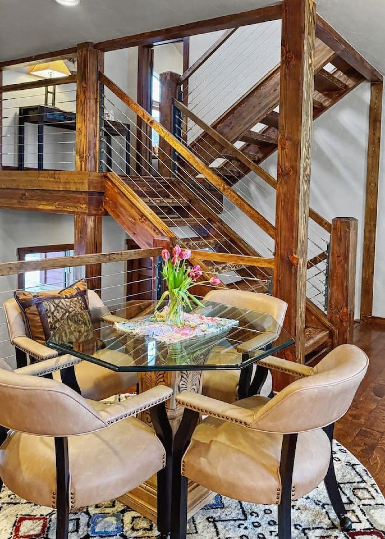 A dining area with a glass table surrounded by leather chairs, a staircase with wooden beams, and a view of the upper floor.