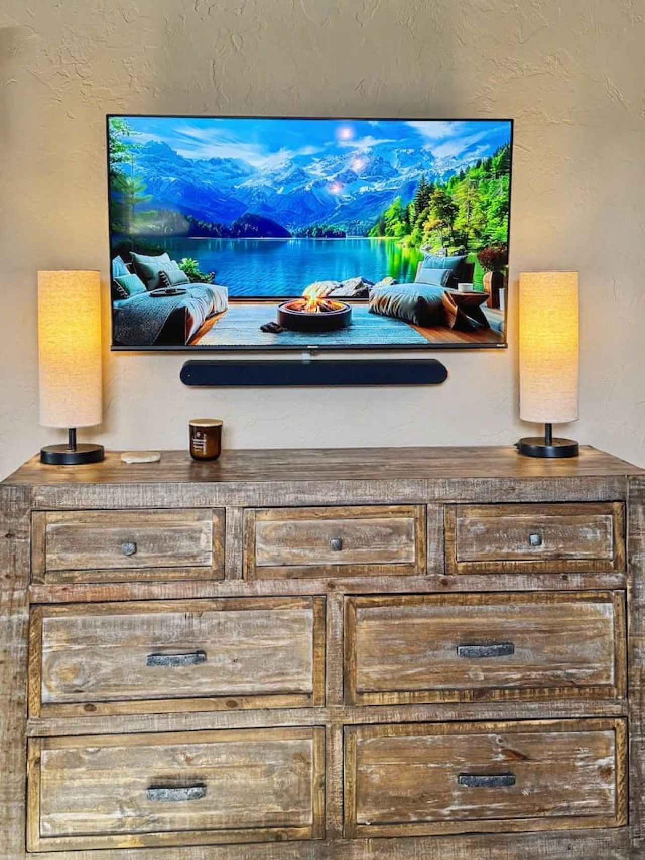 A television mounted on a wall with a scenic landscape image, flanked by two lamps on a wooden cabinet below.