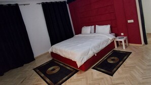 Une chambre d’hôtel avec un lit, une table de chevet, une tête de lit rouge, un parquet en bois à motifs en chevrons et un luminaire au plafond.
