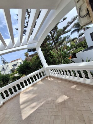 A covered outdoor area with a white railing, paved floor, and views of surrounding buildings and greenery.