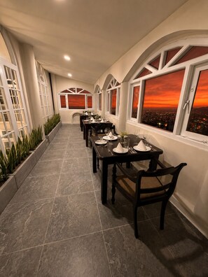 A corridor with a dining area featuring tables set for a meal, large windows with a sunset view, and a tiled floor. - ceylon Breeze kandy (Kandy)