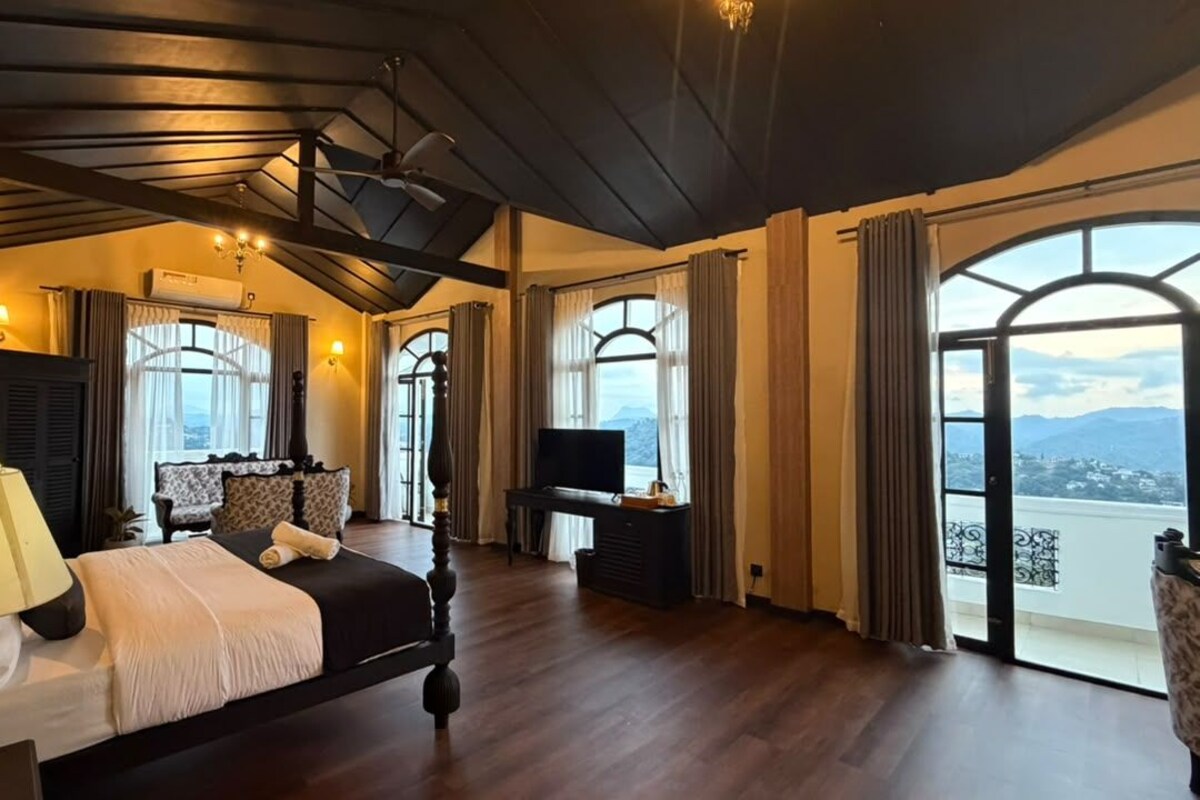 Un dormitorio amplio con una cama grande, un escritorio y una televisión. Varias ventanas ofrecen vistas al exterior.