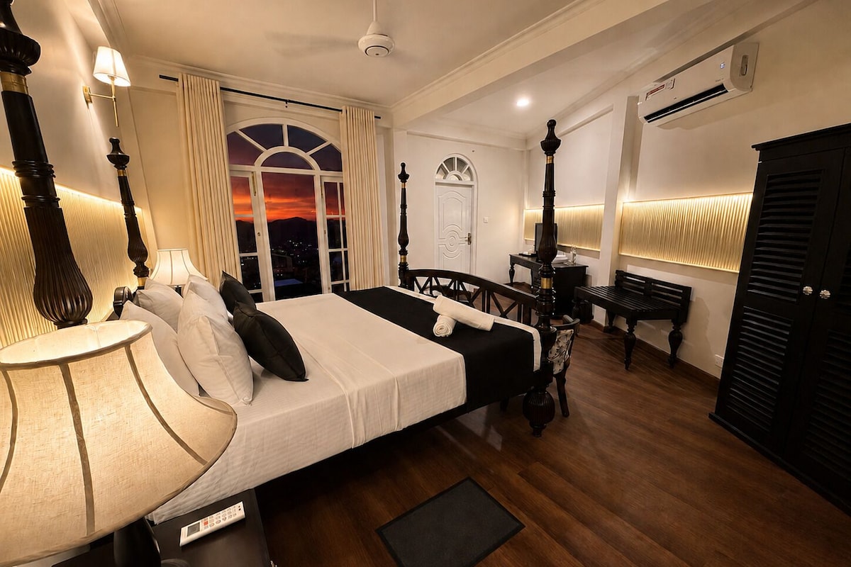 A hotel room with a bed, bedside table, lamp, and a view of the city at sunset.