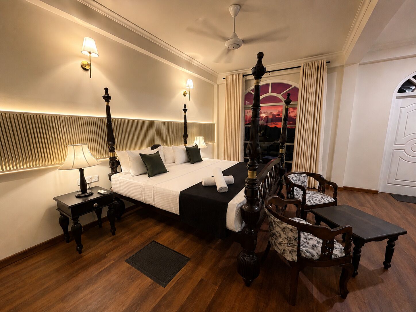 A hotel room with a four-poster bed, a sitting area with a table and chairs, a view of the sunset through a window, and a ceiling fan.