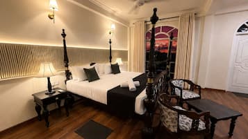 A hotel room with a four-poster bed, a sitting area with a table and chairs, a view of the sunset through a window, and a ceiling fan.