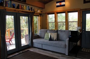 A cozy living room with a grey sofa, a coffee table, and a bookshelf. - Stunning Desert Sanctuary - Tiny House with Mesa Verde Views & Starry Nights (Dolores)