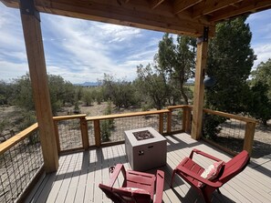 A wooden deck with a fire pit, two red chairs, and a view of a desert landscape with trees and mountains in the distance.