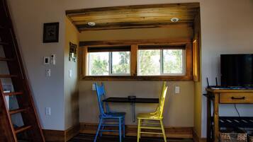 A room with a wooden ceiling, a window, two chairs, a desk, and a television.