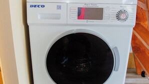 DECO-merkkinen yhdistelmäpesu- ja kuivauskone, joka on sijoitettu huoneen nurkkaan. Huoneessa on puulattia ja valkoinen seinä.