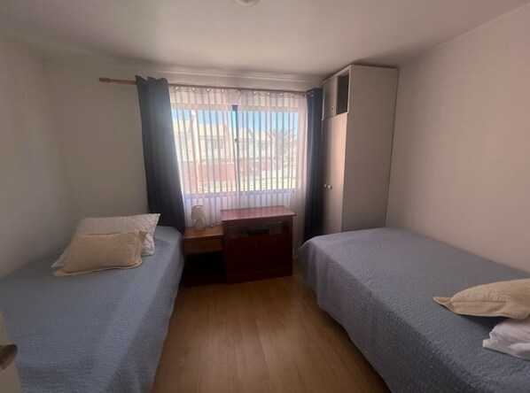 A small, single-room accommodation with two beds, a desk, and a window with curtains.