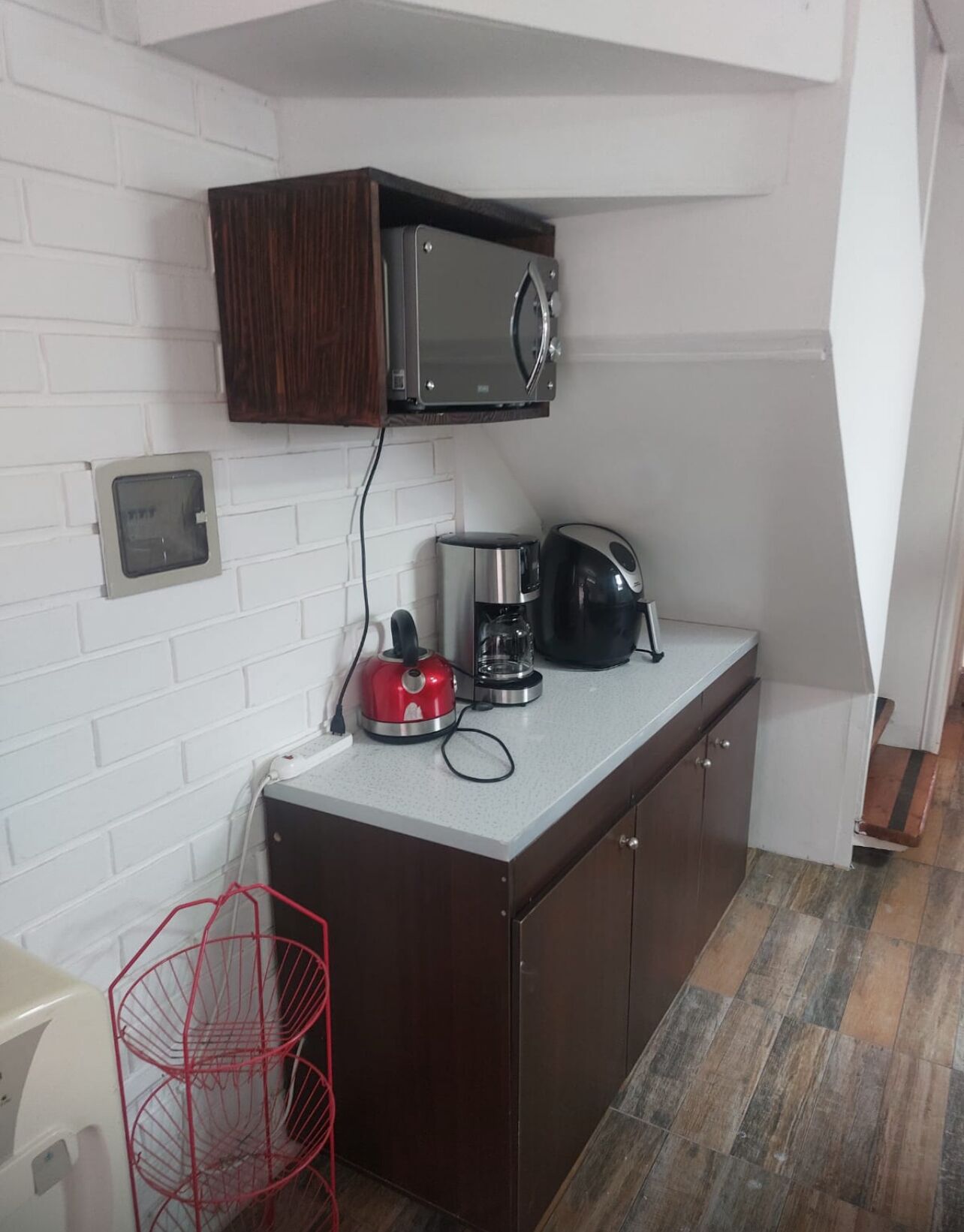 A compact kitchen with a microwave, coffee maker, and kettle on the counter, a red wire basket, and a white brick wall.