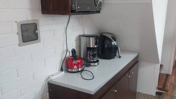 A compact kitchen with a microwave, coffee maker, and kettle on the counter, a red wire basket, and a white brick wall.