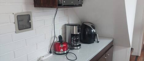 A compact kitchen with a microwave, coffee maker, and kettle on the counter, a red wire basket, and a white brick wall.