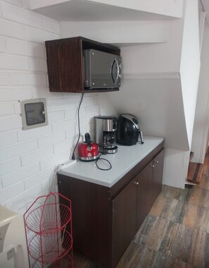 A compact kitchen with a microwave, coffee maker, and kettle on the counter, a red wire basket, and a white brick wall.