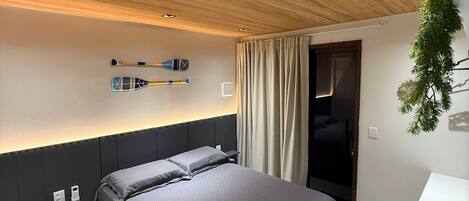 A modern bedroom with a wooden ceiling, a bed with grey bedding, a bedside table, and a desk with a plant.