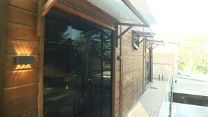 Exterior de madera con puerta de vidrio, una terraza y un muro de ladrillo al fondo.
