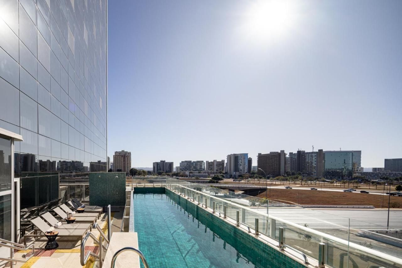 A rooftop swimming pool with lounge chairs, a glass barrier, and a cityscape view.