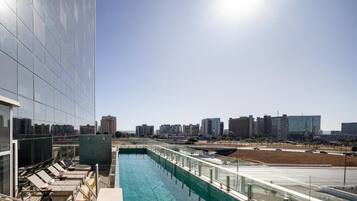 A rooftop swimming pool with lounge chairs, a glass barrier, and a cityscape view.