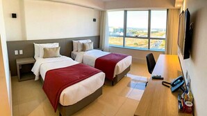 Quarto de hotel com duas camas, uma escrivaninha, uma cadeira, uma TV e uma janela com vista para a cidade.