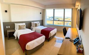 A hotel room with two beds, a desk, a chair, a TV, and a window with a city view. - Flat Particular no Hotel Ramada Brasília (Brasília)