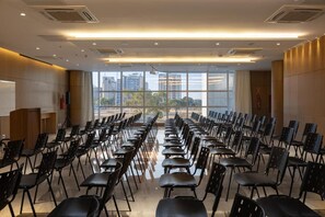 Un salón de conferencias con filas de sillas orientadas hacia un atril, amplias ventanas con vista a la ciudad y un diseño de techo moderno.