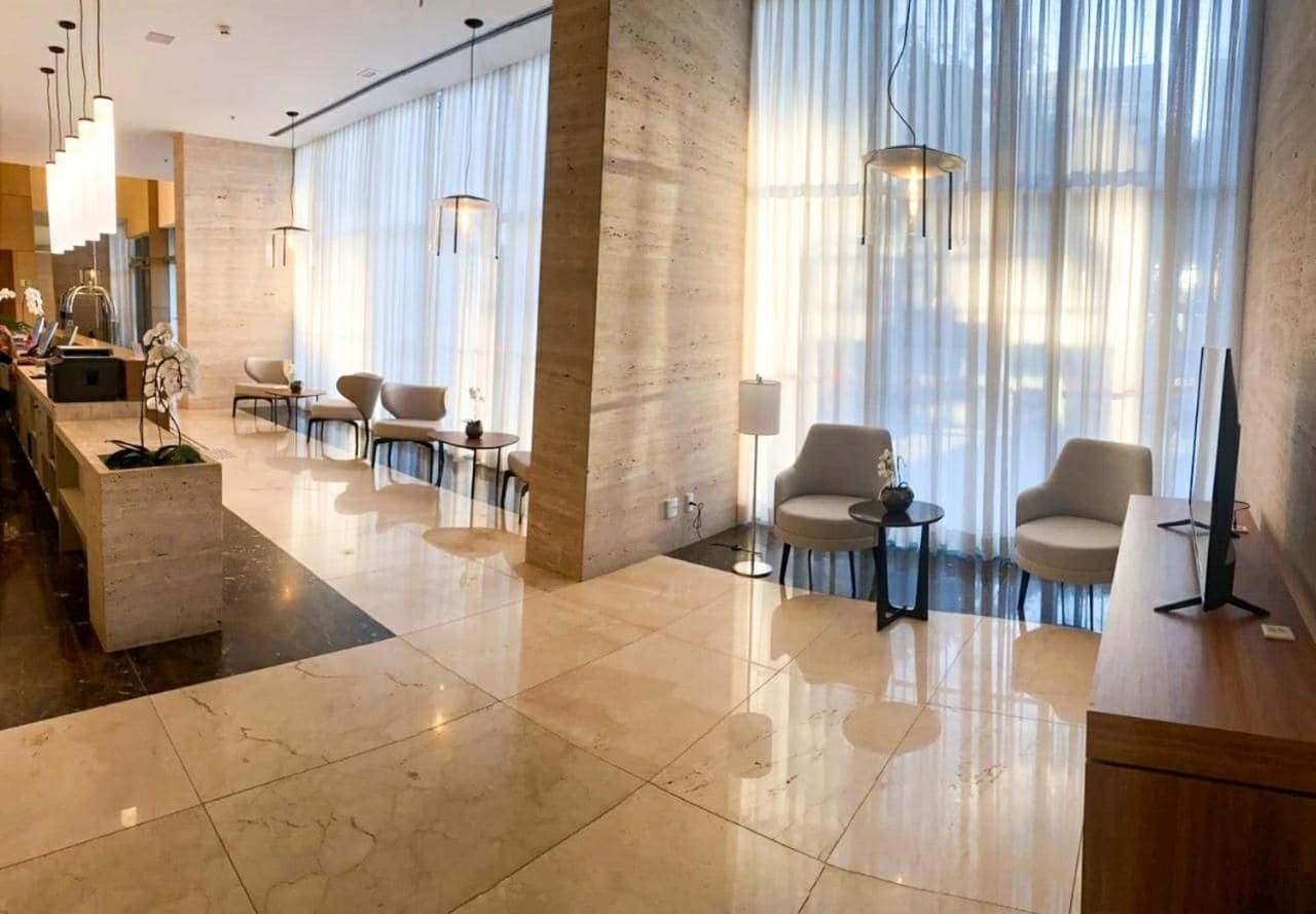 Un moderno vestíbulo de hotel con piso de mármol, zona de estar con sillas y mesas, un mostrador de recepción y ventanas amplias con cortinas.
