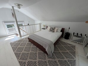 A bedroom with a sloped ceiling, a bed with patterned bedding, a nightstand, a lamp, and a window with a view of greenery. - The Beech Street Bungalow-  Hottub + Pedal Boat (Smallwood)