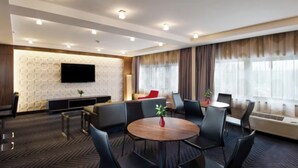 Una camera d'albergo moderna con un tavolo da pranzo rotondo, posti a sedere confortevoli, una TV a schermo piatto montata su una parete strutturata e ampie finestre con tende.