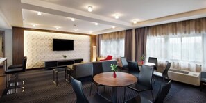Habitación de hotel moderna con una mesa de comedor redonda, asientos cómodos, un televisor de pantalla plana montado en una pared texturizada y ventanas amplias con cortinas.