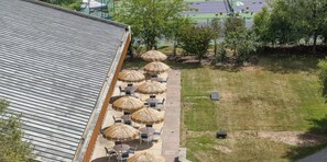 Un edificio con techo inclinado, mesas al aire libre con sombrillas y un área verde con árboles.