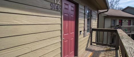 A wooden house with a red door, a number '12 9145' on the wall, and a small porch with a wooden railing.