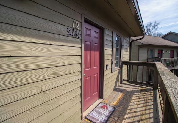 A wooden house with a red door, a number '12 9145' on the wall, and a small porch with a wooden railing.