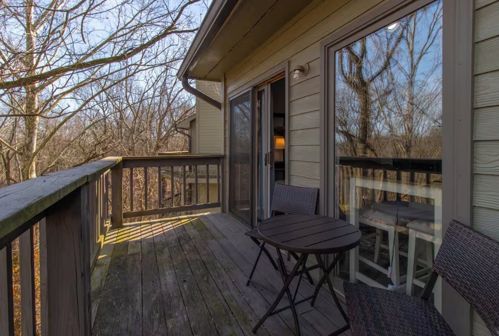 A wooden deck with a table and chairs, a glass door leading to a room, and a view of trees.