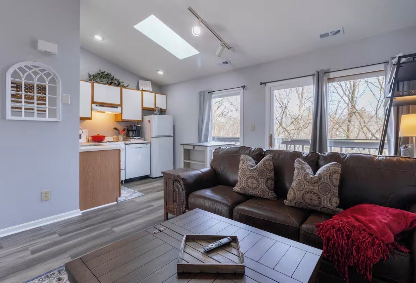 A modern living room with a brown leather sofa, a wooden coffee table, and a kitchen area with white cabinets and a red appliance.