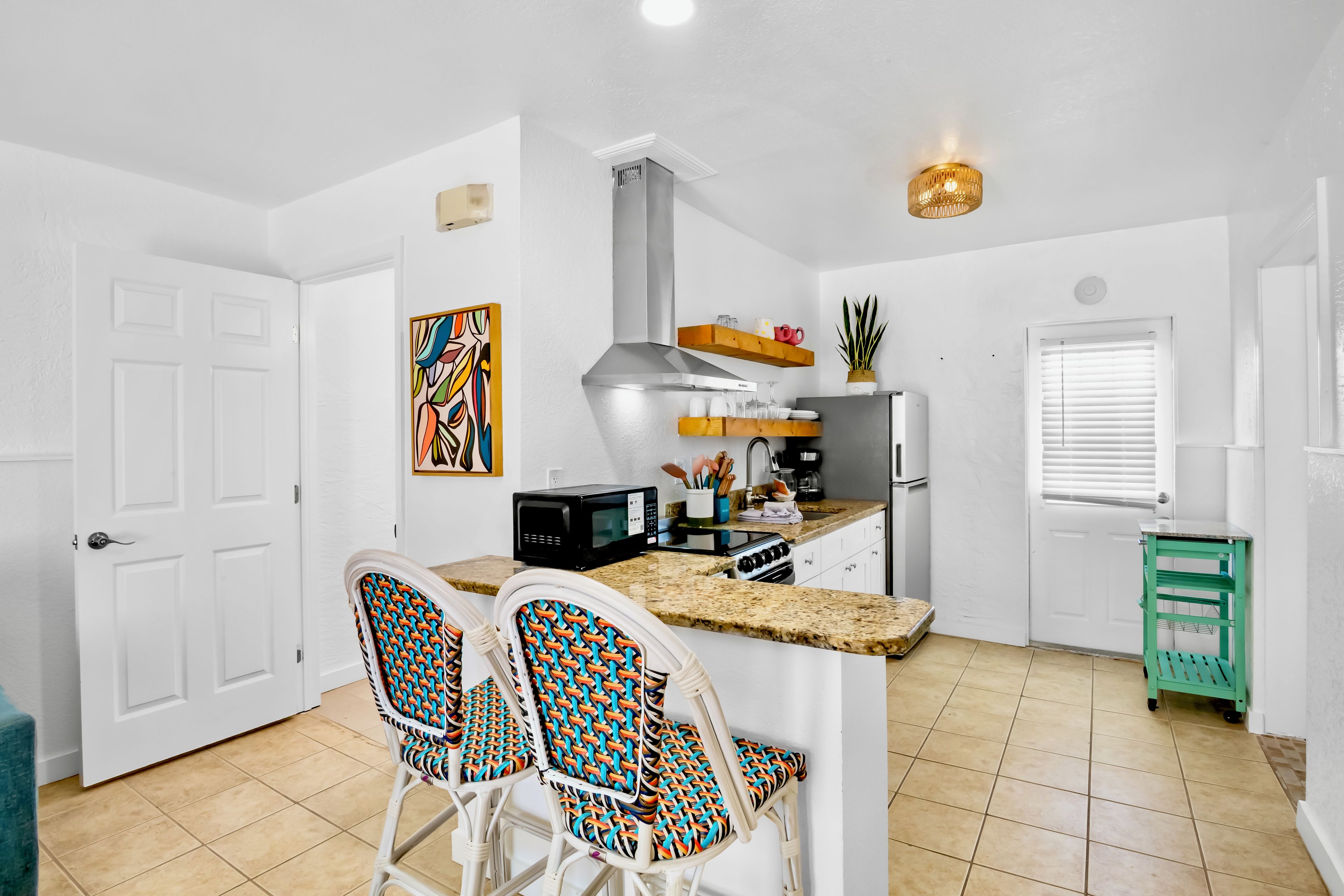 Cocina compacta con una isla central, muebles blancos y una zona de comedor con sillas de colores.
