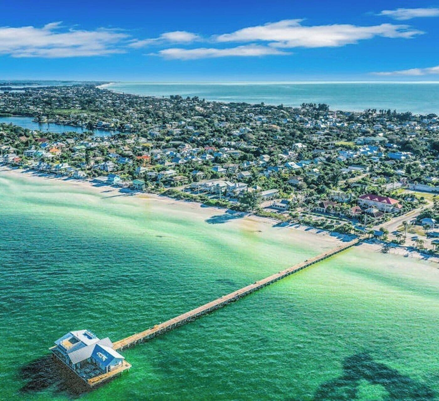 Vista aérea de una zona costera con un muelle largo que se extiende hacia el agua, rodeado de edificios residenciales y un cielo azul despejado.