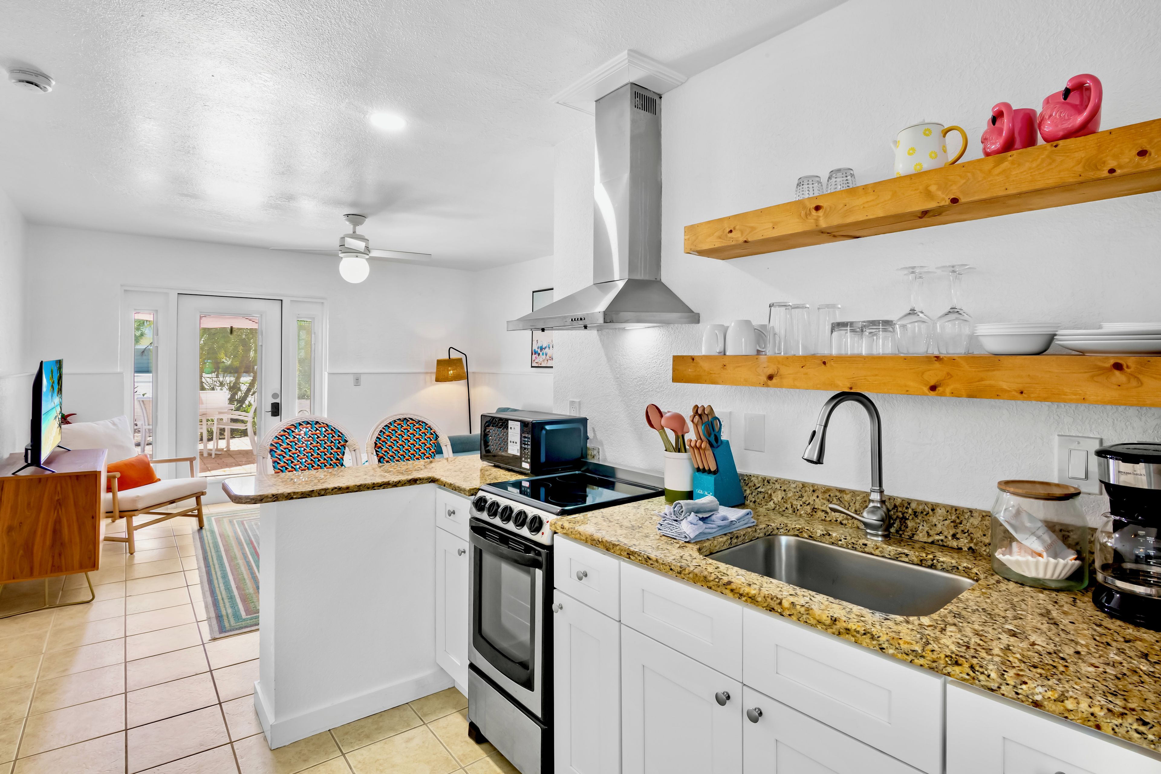 A compact kitchen with white cabinetry, a stove, microwave, and a sink. There are wooden shelves above the counter holding kitchenware.