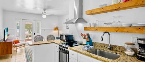 Une cuisine compacte avec des meubles blancs, une cuisinière, un four à micro-ondes et un évier. Des étagères en bois sont fixées au-dessus du plan de travail et servent à ranger les ustensiles de cuisine.