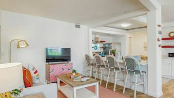 Una sala de estar moderna con una televisión, un mueble rosa y una zona de comedor con sillas blancas.