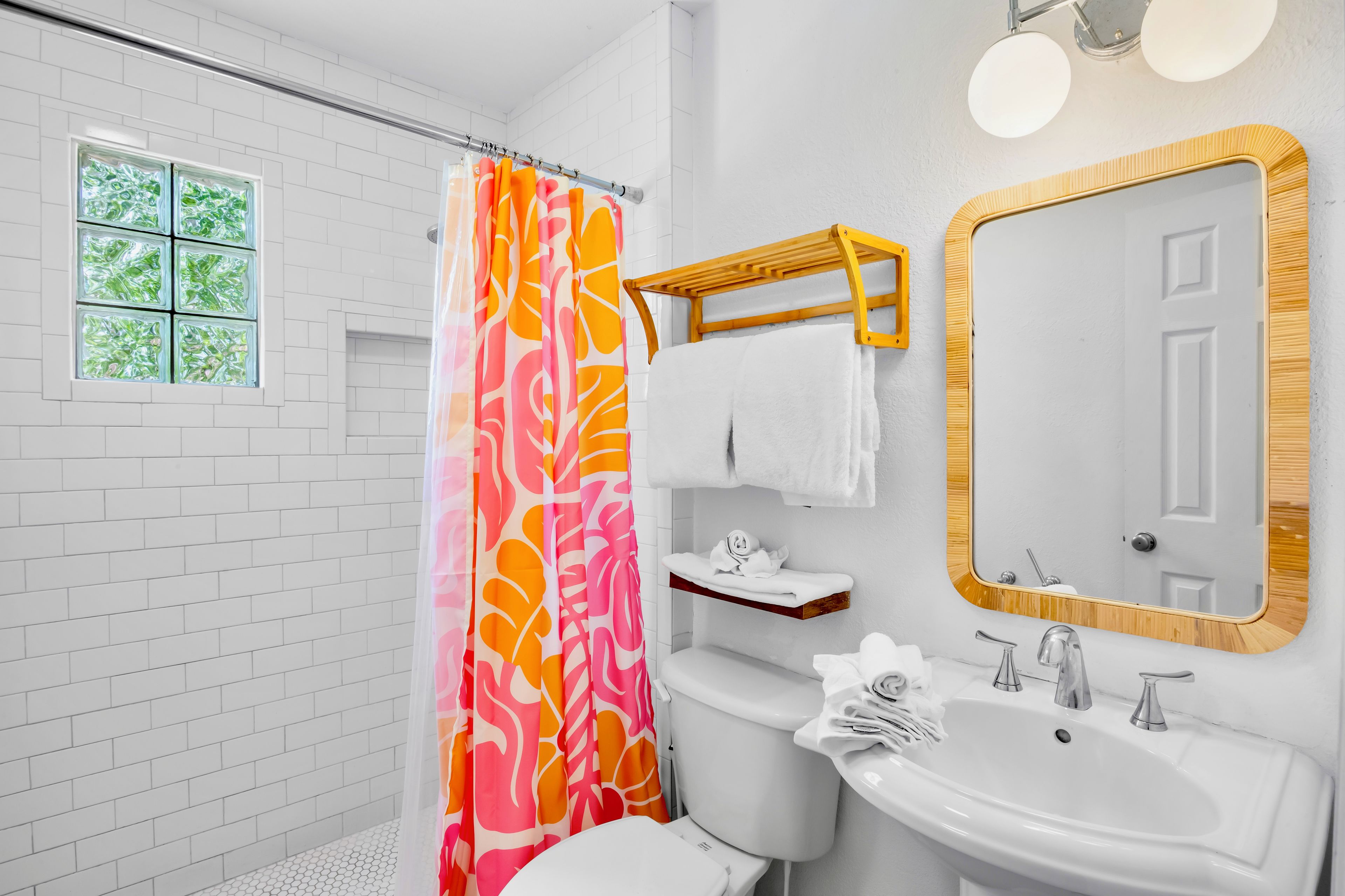 A bathroom with a white tiled wall, a shower with a colorful curtain, a toilet, a sink with a mirror, and a window with a view of greenery.