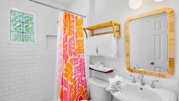 A bathroom with a white tiled wall, a shower with a colorful curtain, a toilet, a sink with a mirror, and a window with a view of greenery.