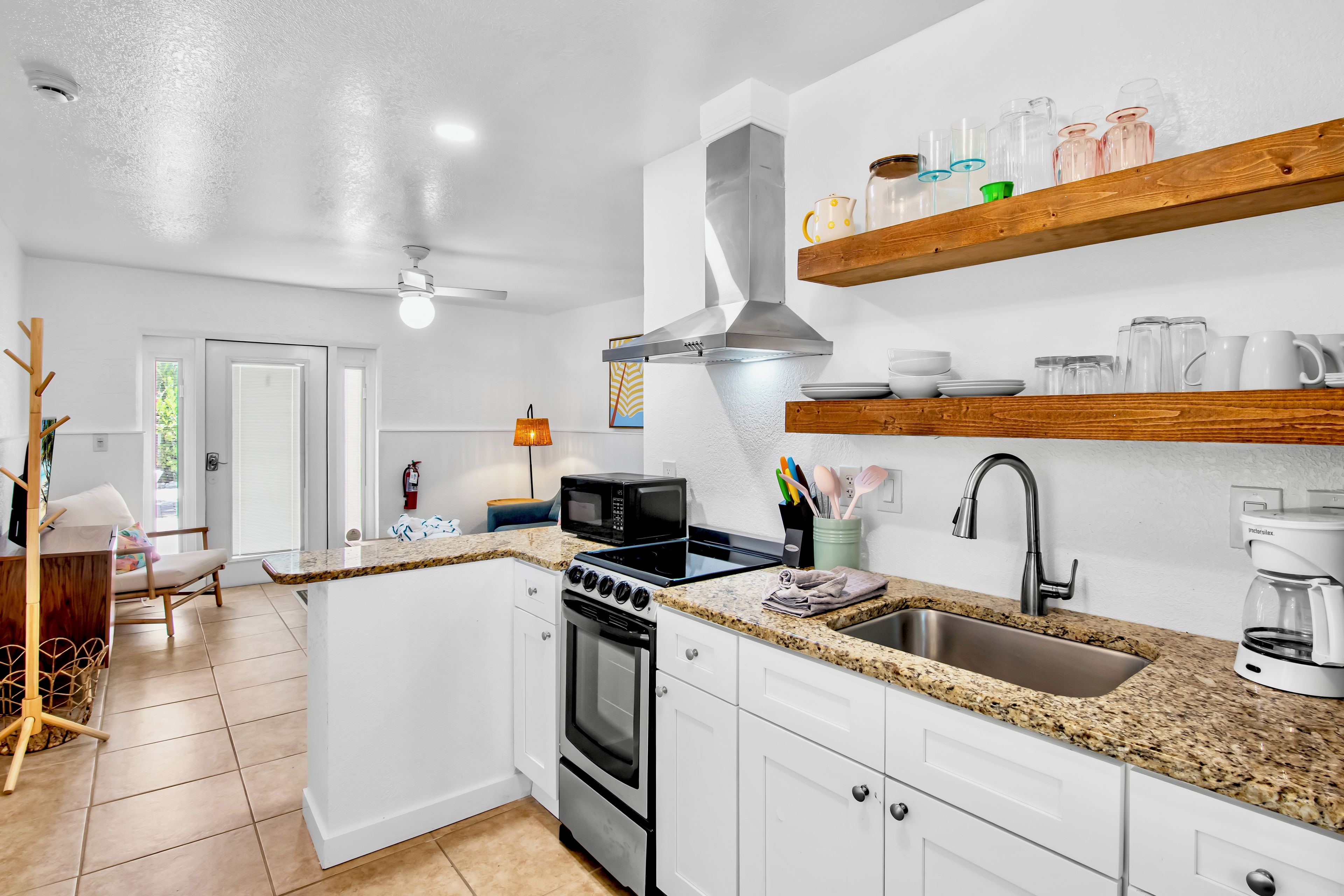 Una cocina moderna con armarios blancos, encimera de granito, pileta de acero inoxidable, horno empotrado y microondas. Hay estantes de madera sobre la encimera con cristalería.