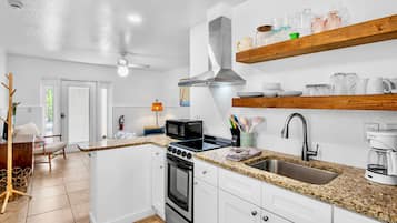 Una cocina moderna con armarios blancos, encimera de granito, pileta de acero inoxidable, horno empotrado y microondas. Hay estantes de madera sobre la encimera con cristalería.