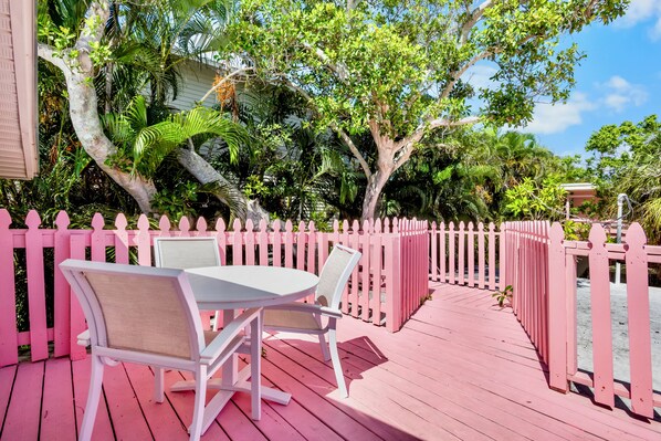 A pink wooden deck with a table and chairs, surrounded by a pink fence and lush greenery.