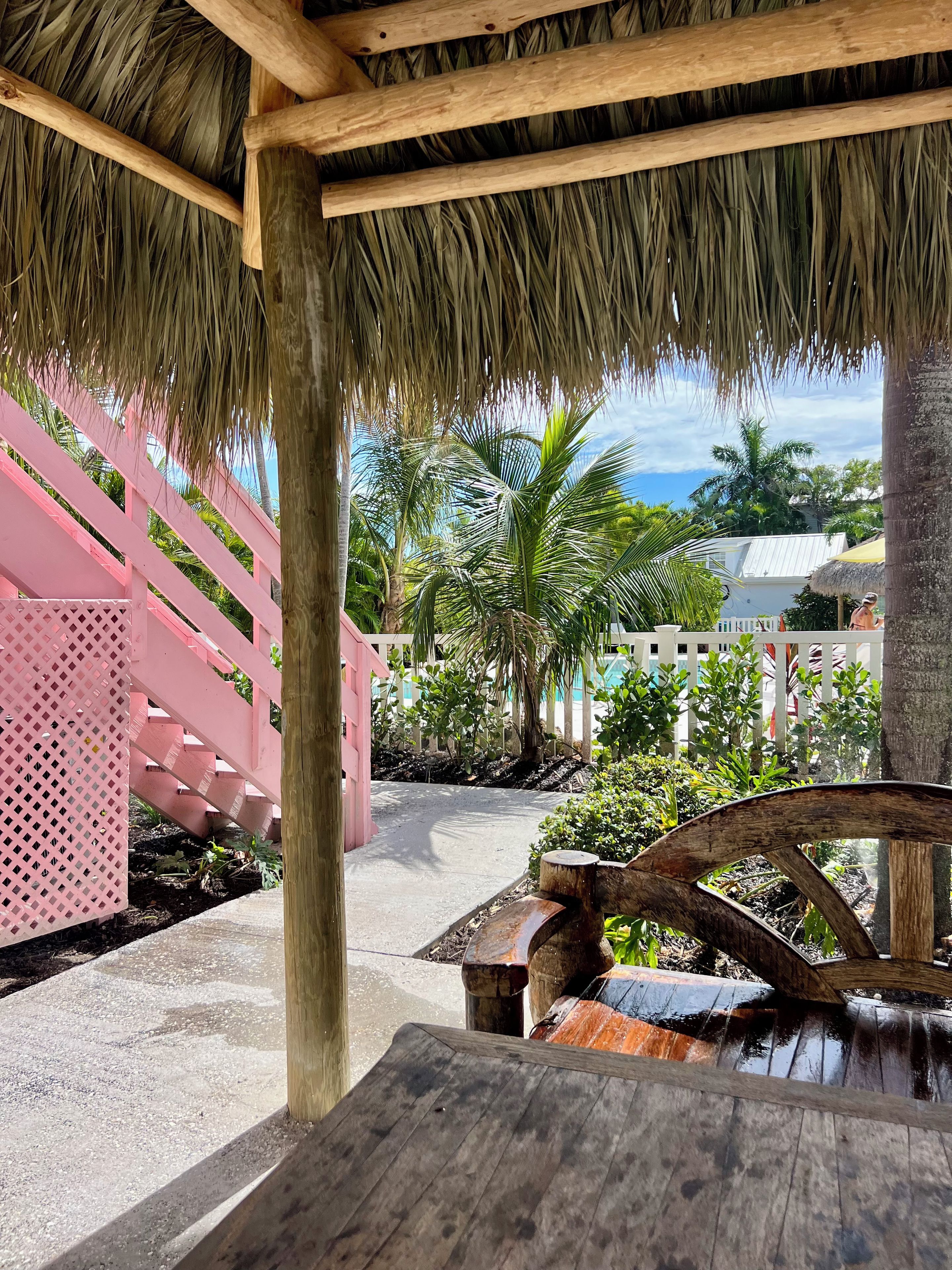 A covered outdoor area with a wooden table, a thatched roof, and a pink fence.