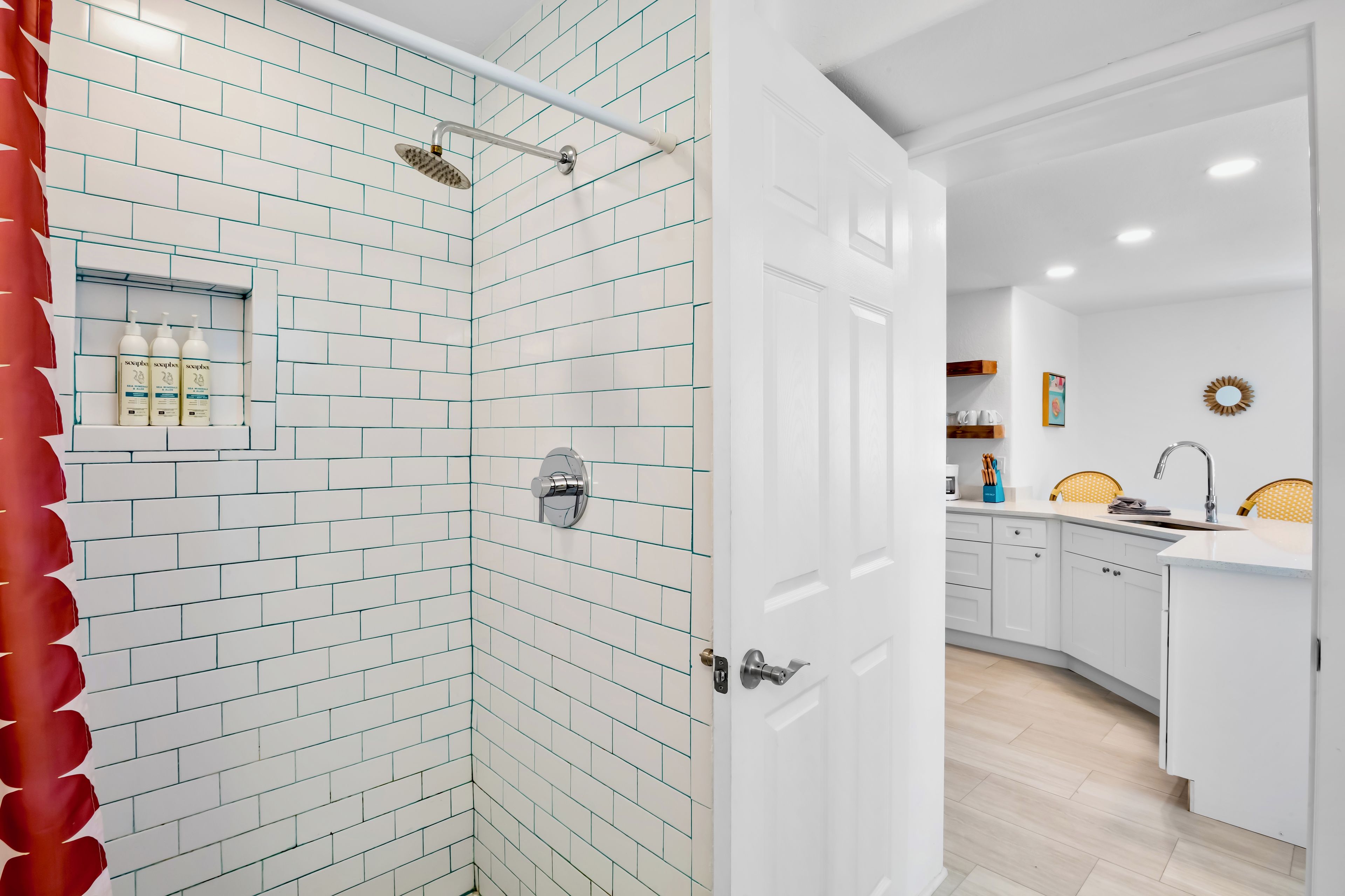 Un baño moderno con azulejos blancos tipo metro, una ducha con cabezal de ducha manual y una puerta blanca que da a una zona de cocina.